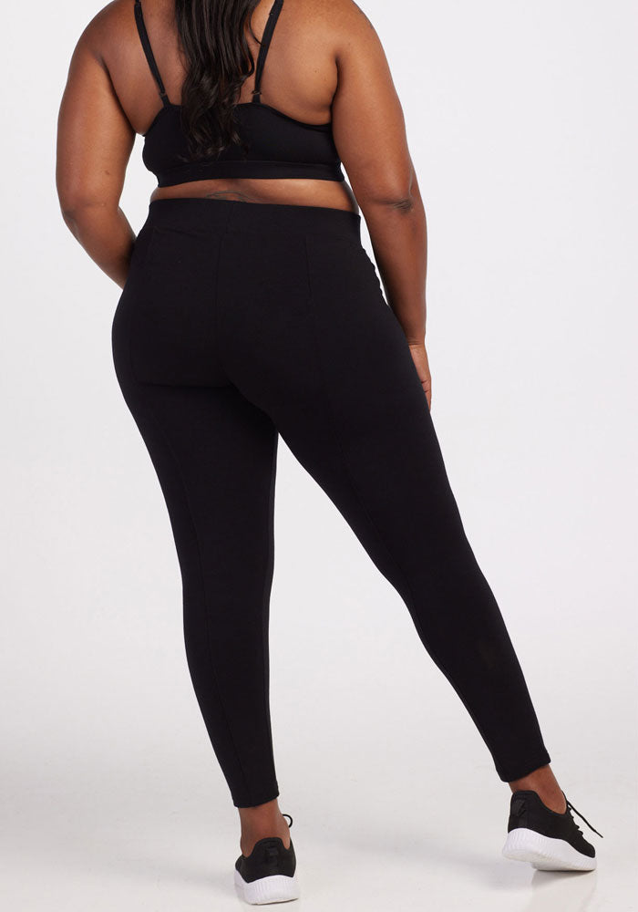 Rear view of a person wearing Stella Leggings in black from Woolx and a black sports bra. The person is standing with one leg slightly bent, looking to the left while sporting black and white sneakers. The plain white background perfectly highlights the cold weather baselayers.