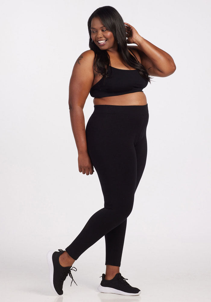 A woman with long dark hair smiles while wearing a black sports bra, Stella Leggings in Black from Woolx, and black sneakers. She is striking a pose with one hand touching her hair and the other resting on her hip, standing against a plain white background.