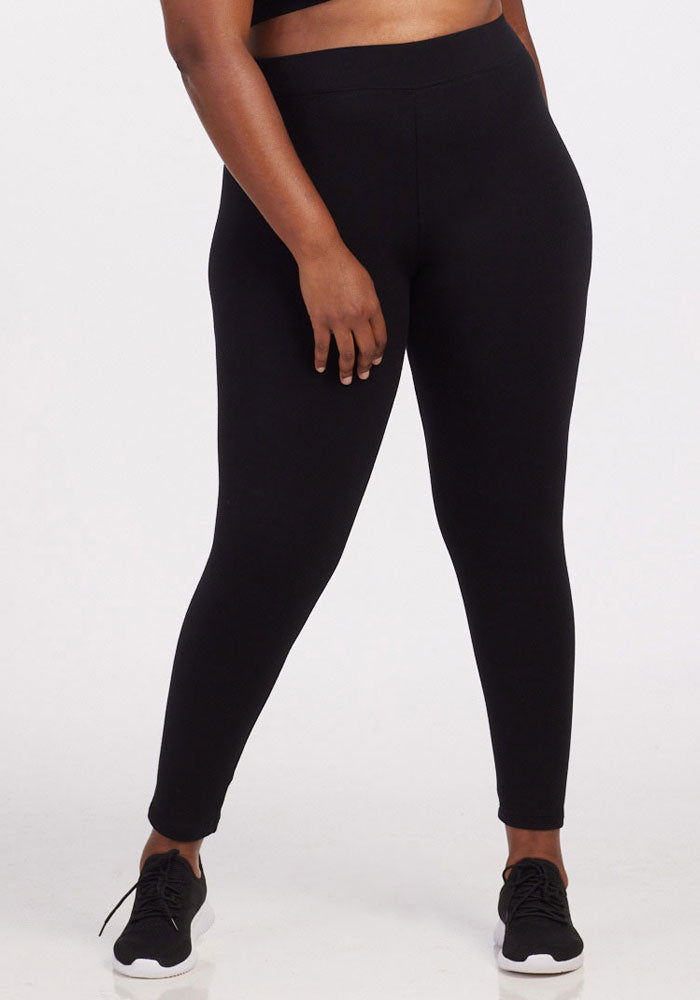 A person wearing the Stella Tall in black by Woolx and black athletic shoes with white soles stands facing forward. Only the body from the waist down is visible in the image. The person’s right hand rests along the side of their thigh, showcasing the sleek design of these DuraLite fabric leggings. 