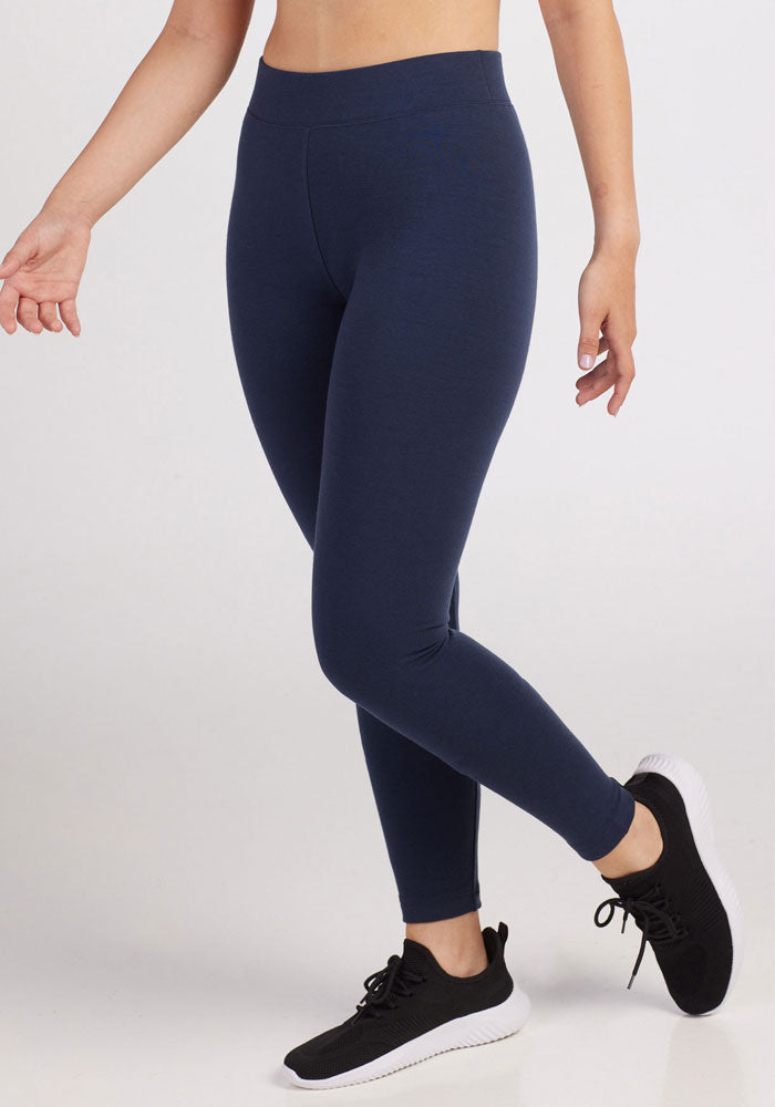 A person wearing deep navy Stella Leggings by Woolx and black athletic shoes stands with one leg slightly lifted and arms relaxed by their sides against a plain background. Their midsection and legs are in focus, highlighting the fit of the cold weather baselayer.