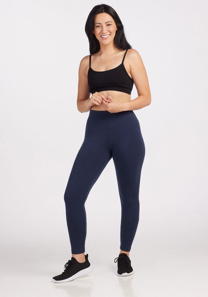 A woman with long, dark hair is standing against a plain background. She is wearing a black sports bra, deep navy Stella leggings from Woolx Duralite Fabric, and black athletic shoes. She is smiling and has her hands clasped in front of her.