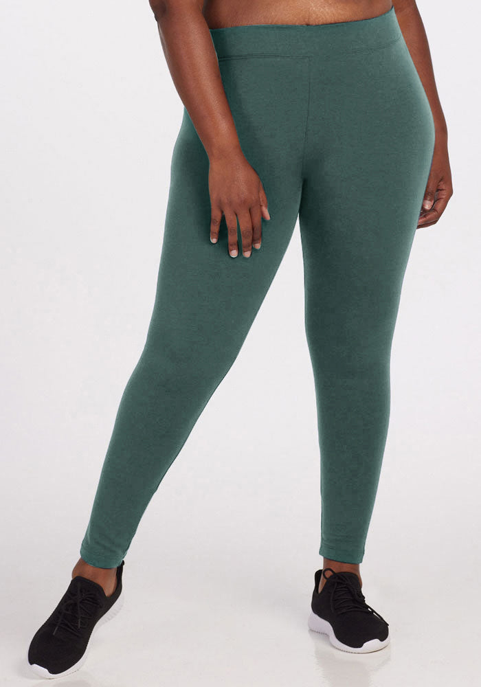 A person wearing dark green Stella Leggings by Woolx and black sneakers stands against a plain white background. The photo is cropped to show the individual from their stomach to their feet, with their right hand resting on their thigh. 