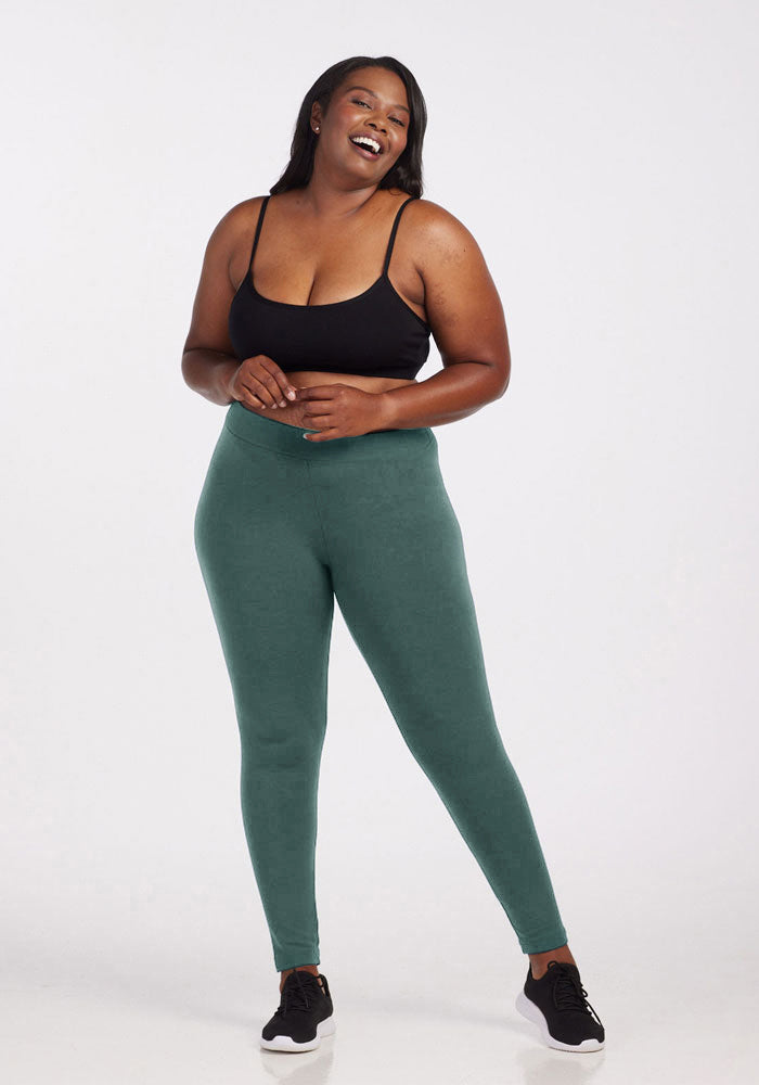 A person is standing and laughing, wearing a black sports bra, Woolx green Stella Leggings, and black sneakers. They have long, dark hair and are positioned against a plain, light-colored background.