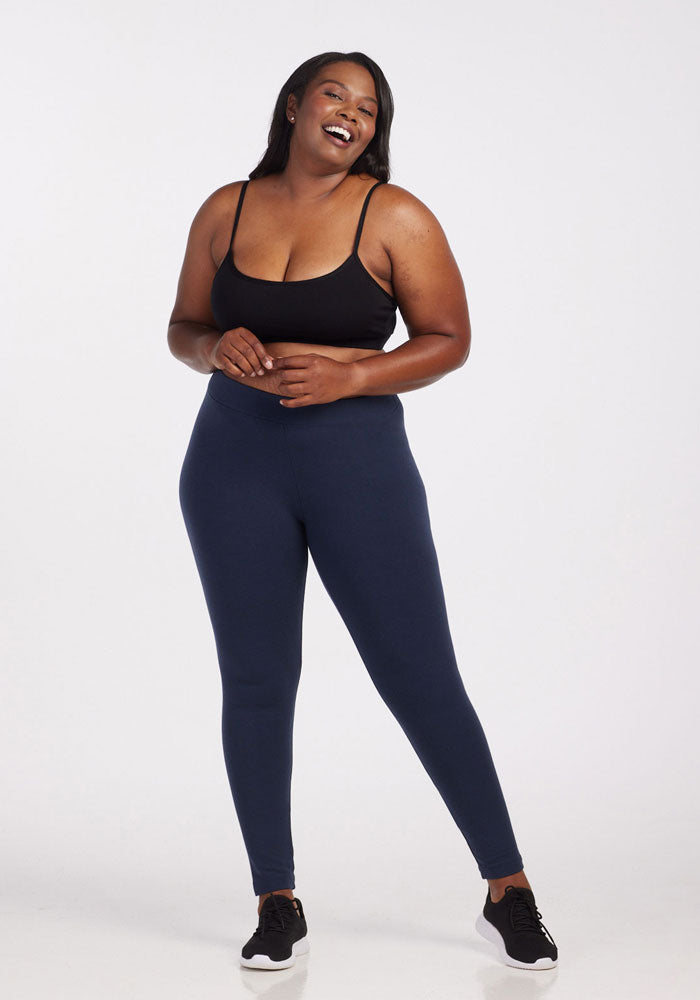 A woman smiles and laughs while posing in front of a plain white background. She is wearing a black sports bra, deep navy Stella Tall leggings by Woolx, and black athletic shoes. The setting appears to be a studio.