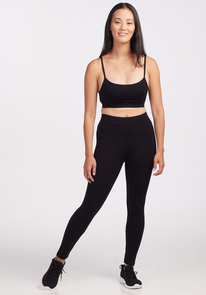 A young woman with long dark hair stands smiling against a plain background. She is wearing the Stella Petite - Black sports bra by Woolx, comfortable fit Merino wool leggings, and black sneakers with white soles.