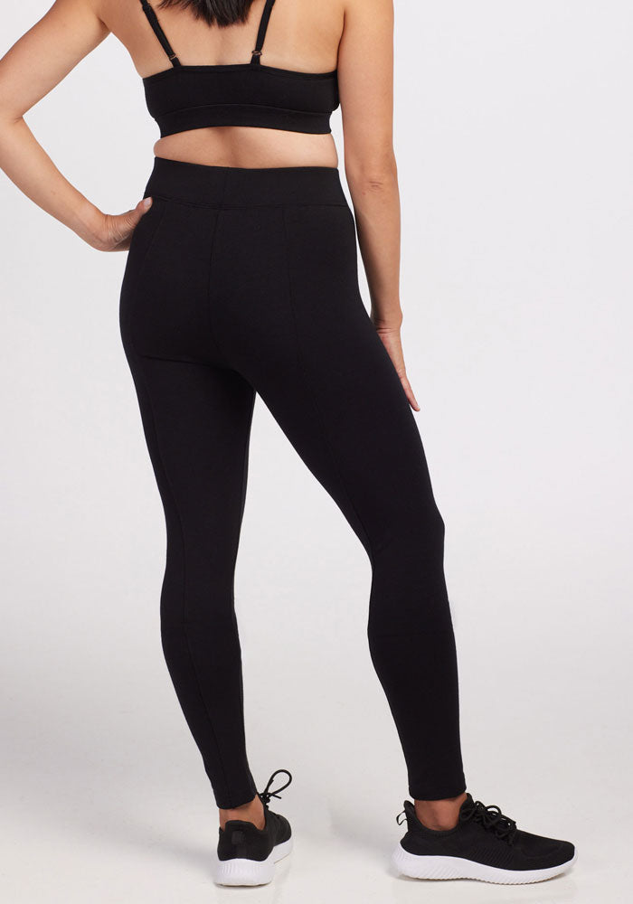 A person wearing the Woolx Stella Petite - Black sports bra, comfortable fit black high-waisted leggings, and black athletic shoes stands facing away, with their left hand resting on their hip and right arm at their side. They are standing on a plain, light-colored background.