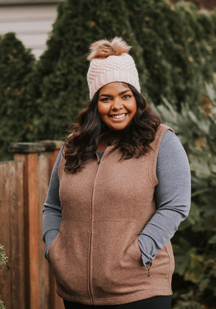 A person wearing a brown zip-up vest over a blue long-sleeve shirt and the Woolx Kaylee Beanie in rose, featuring a chunky knit with a faux fur pom-pom, stands outdoors smiling. Greenery and a wooden fence provide the background.