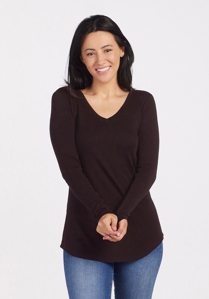 A woman with long, dark hair smiles at the camera. She is wearing a long-sleeved Woolx Eva Tunic in French Roast and blue jeans, embodying everyday street style. Her hands are clasped together in front of her, set against a plain white background.