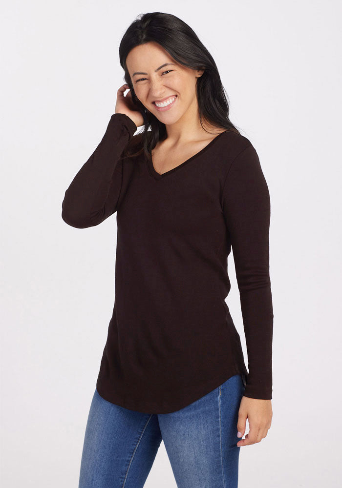 A woman with long dark hair, wearing the Woolx Eva Tunic in French Roast and blue jeans, is standing and smiling. She has one hand raised to her ear and appears to be in a cheerful and relaxed mood. Her street style against the plain white background exudes effortless charm.