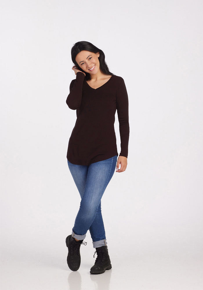 A person with long, dark hair is standing and smiling while tilting their head slightly. They are wearing a long-sleeved French Roast Eva Tunic by Woolx, paired with blue jeans and black boots. The ensemble exudes a casual street style against a plain white background.