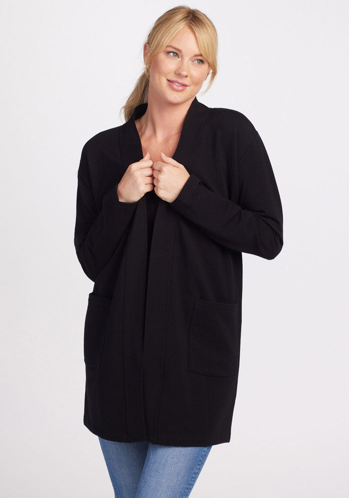 A person with blond hair pulled back is wearing the Kinsley Cardigan - Black by Woolx paired with blue jeans. They are standing against a plain white background and slightly smiling while holding the cardigan's front edges near their collar, enjoying its heavyweight warmth and convenient side pockets.