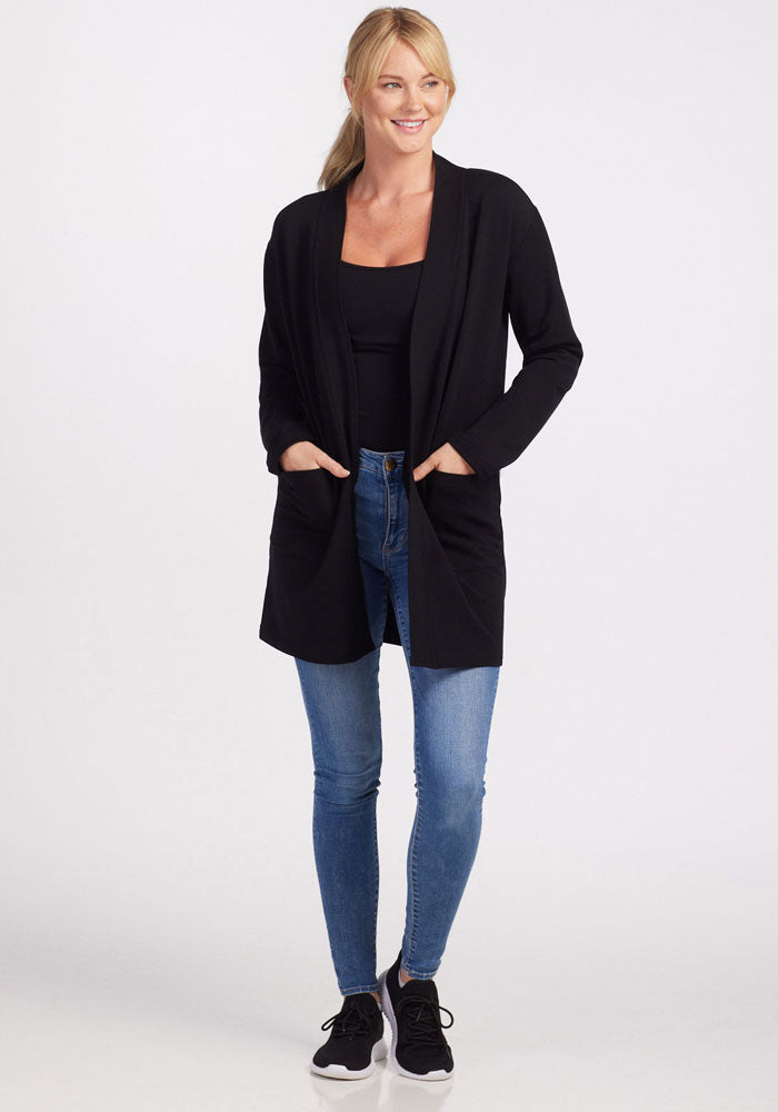 A woman stands smiling in the Woolx Kinsley Cardigan in black, paired with a black top, blue jeans, and black sneakers against a plain white background. She has blonde hair tied back and her hands are in the cardigan's side pockets.