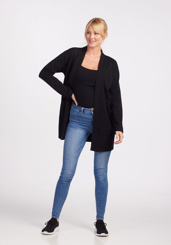 A person with blonde hair, tied back in a ponytail, is standing and smiling. They are wearing a Woolx Kinsley Cardigan in black over a black top, blue skinny jeans, and black sneakers with white soles. One hand is resting on their hip as they stand against a plain white background.