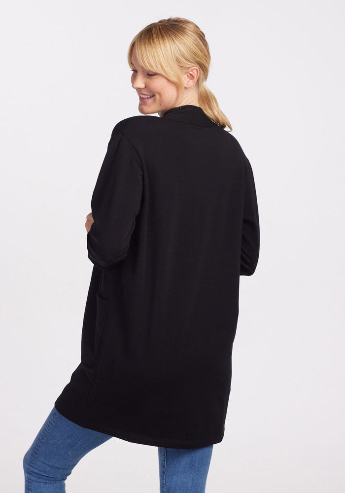 A person with blonde hair pulled back into a ponytail, wearing a long black Kinsley Cardigan by Woolx with side pockets and blue jeans, is smiling and looking over their shoulder. The background is plain white.