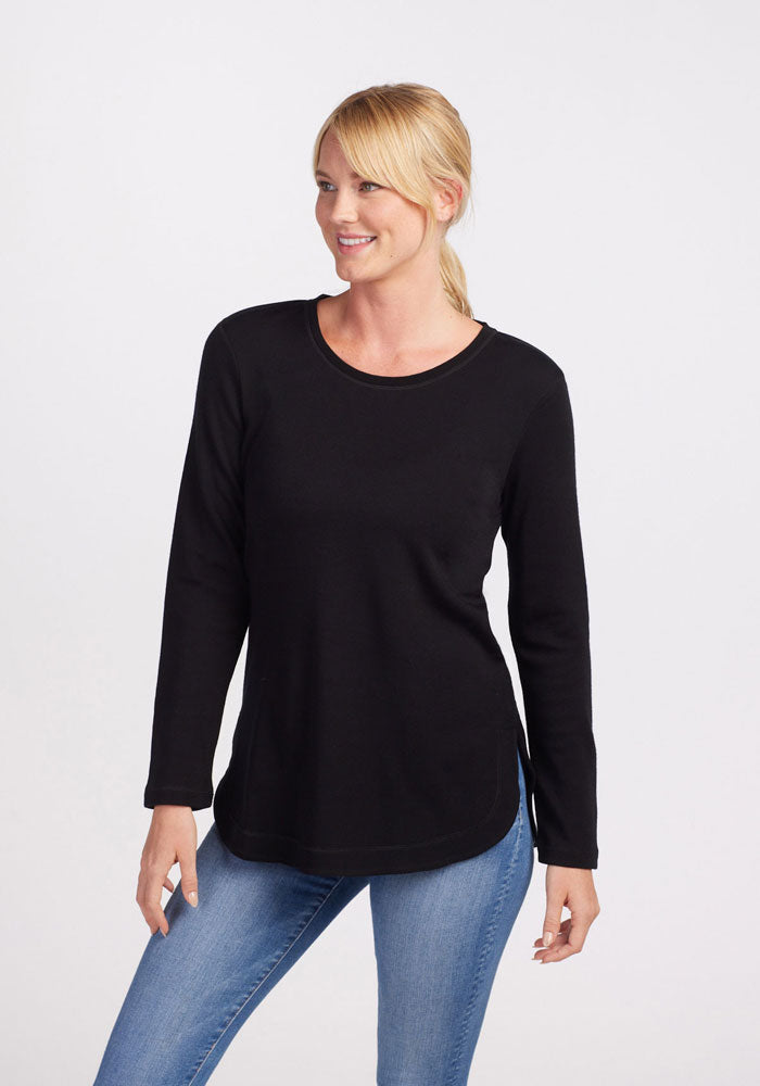 A person with blonde hair is smiling and wearing the ultra-cozy, long-sleeved Blair Tunic - Black by Woolx, paired with light blue jeans. The individual stands against a plain white background with their arms relaxed by their sides.