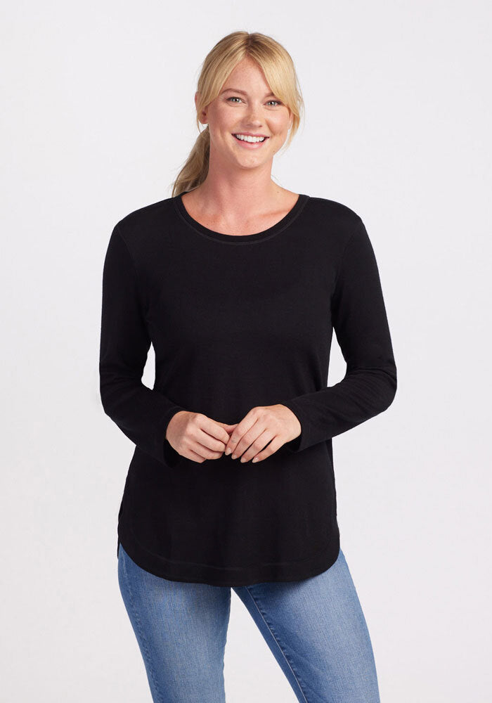 A woman with blonde hair tied back in a ponytail is smiling at the camera. She is wearing the Woolx Blair Tunic - Black, an ultra-cozy, long-sleeve top made from Australian Merino Wool, paired with light blue jeans. Her hands are clasped together in front of her against a plain white background. 