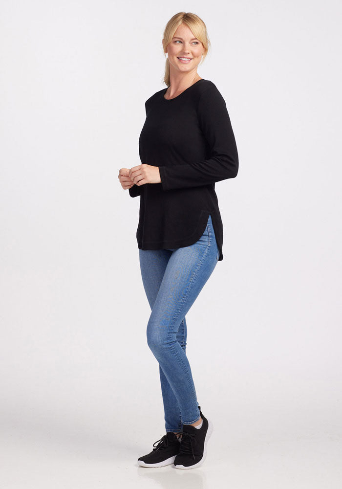 A woman stands and poses against a plain background, smiling. She is wearing the black Blair Tunic from Woolx, paired with light blue jeans and black sneakers. She appears relaxed with her hands clasped gently in front. Her blonde hair is tied back in a loose ponytail.