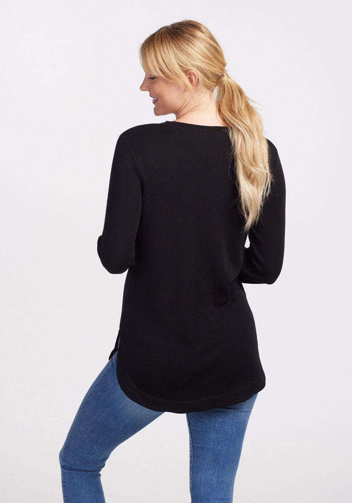 A person with long, blonde hair tied in a ponytail is seen from behind, wearing the Blair Tunic - Black by Woolx made from ultra-cozy Australian Merino Wool and blue jeans. The background is plain and light-colored.