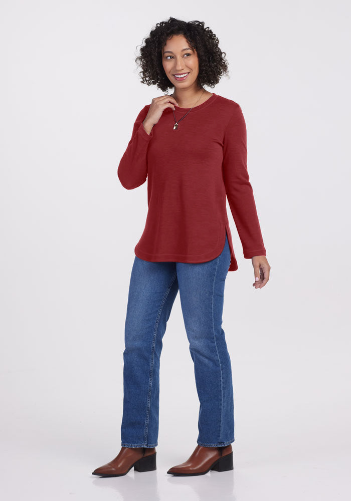 A person with curly hair is posing and smiling, wearing a long-sleeved red Blair Tunic – Sweet Pepper from Woolx, blue jeans, and brown ankle boots. They have one hand lightly touching their neck and the other relaxed by their side. The background is plain white.