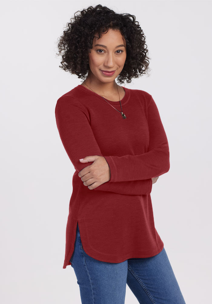 A person with curly black hair, wearing a long-sleeved Blair Tunic in Sweet Pepper from Woolx and blue jeans, stands with their arms crossed against a plain white background. They are smiling softly and wearing a small pendant necklace.