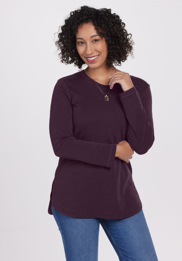 A curly-haired individual smiles while wearing the Woolx Blair Tunic in Deep Plum and blue jeans. A necklace adorns their neck as they pose against a light background, one hand on their hip and the other gently touching their neck.