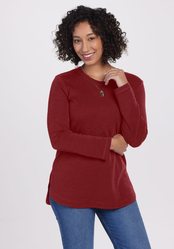 A person with curly hair is smiling while wearing a long-sleeve, maroon Blair Tunic - Sweet Pepper from Woolx, paired with blue jeans. They are standing against a plain white background, with one hand touching their hair and the other arm resting by their side.