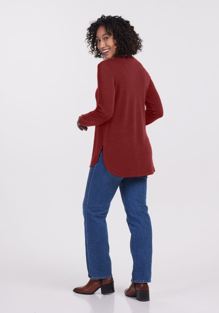 A person with curly hair is smiling and looking back, wearing the Blair Tunic in Sweet Pepper from Woolx, blue jeans, and brown boots. The background is plain white.