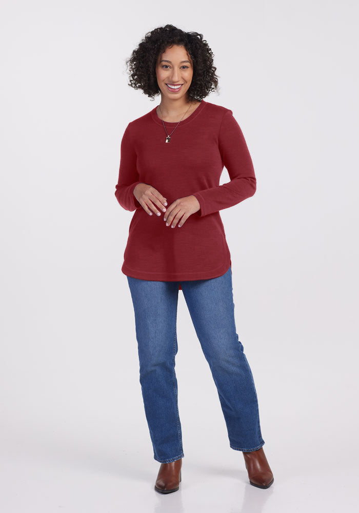 A person with curly hair stands smiling against a plain backdrop. They are wearing a long-sleeved, red Blair Tunic - Sweet Pepper by Woolx, blue jeans, and brown boots. Their hands are gently clasped in front, and they appear relaxed and friendly.