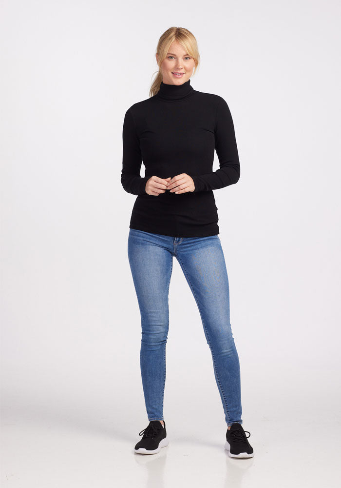 A woman with blonde hair stands confidently, wearing the Woolx Sage Turtleneck in black, blue skinny jeans, and black sneakers. She poses against a plain white background, hands loosely clasped in front of her.