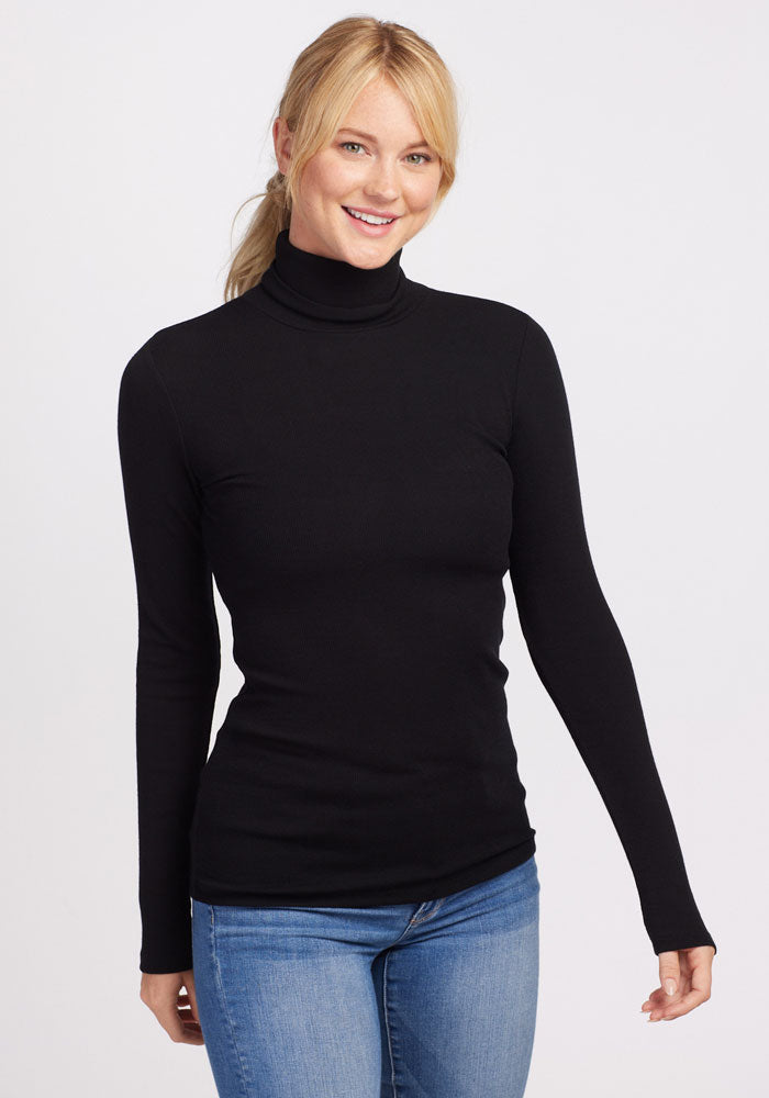 A person with blonde hair tied back is smiling and wearing a Woolx Sage Turtleneck in black along with blue jeans. They are standing against a plain white background.