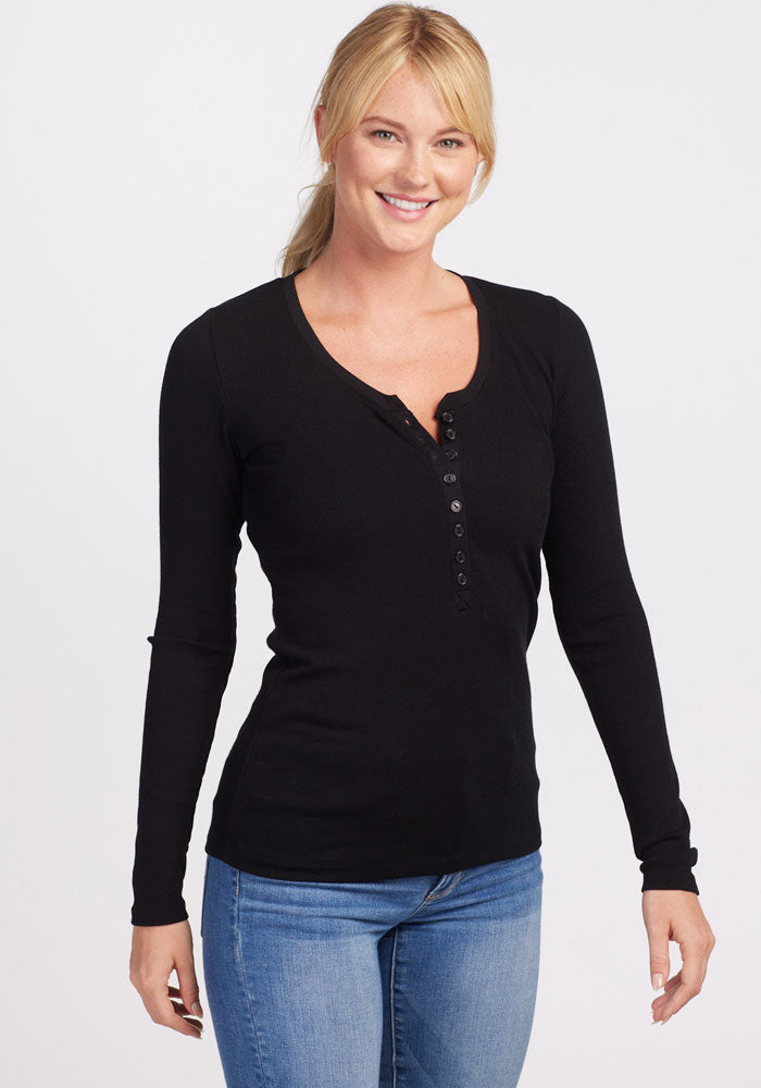 A smiling person with blonde hair is wearing the Reese Ribbed Top - Black from Woolx, paired with blue jeans. They are standing against a plain white background.