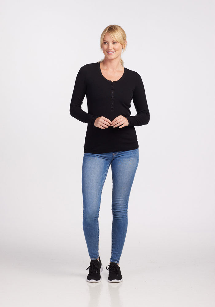 A person with blonde hair is standing against a plain white background. They are wearing a fitted Reese Ribbed Top in Black from Woolx, blue jeans, and black sneakers. Their hands are loosely clasped in front, and they have a slight smile on their face.