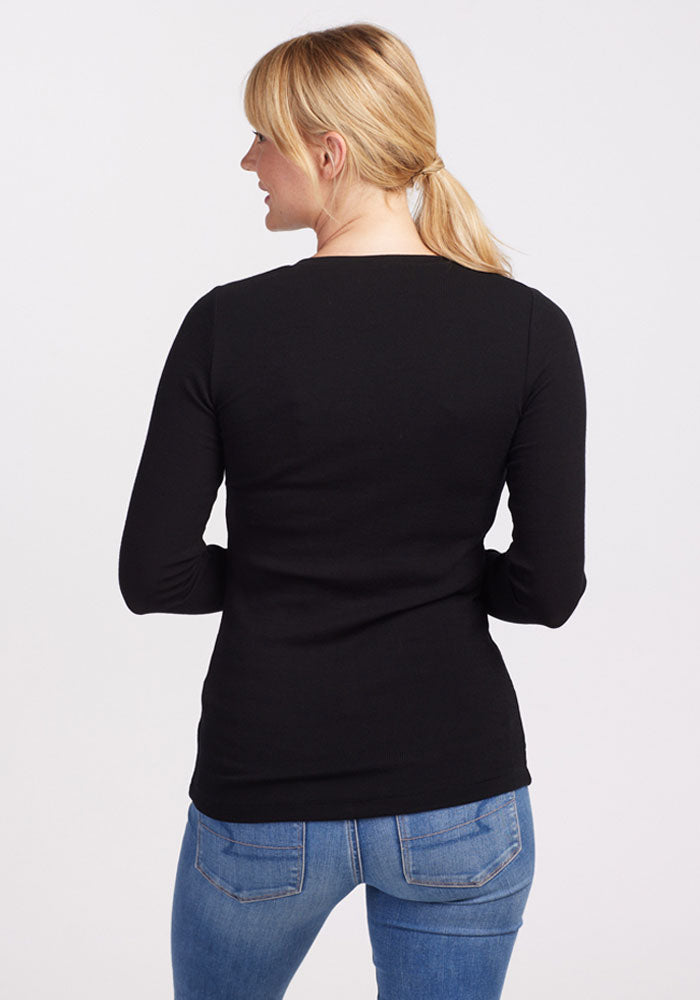 A person with blonde hair tied in a low ponytail is seen from the back, wearing a black Reese Ribbed Top by Woolx and blue jeans. The background is plain and light-colored.