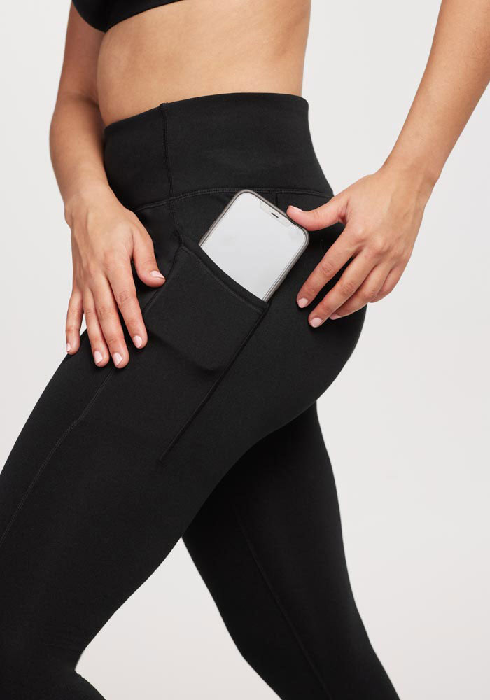 A person in Merino Flex™ MCKENNA PETITE leggings and a sports top slides a smartphone into the handy side pocket, with the plain white background highlighting the functionality of these sporty, Woolx leggings infused with Merino wool.