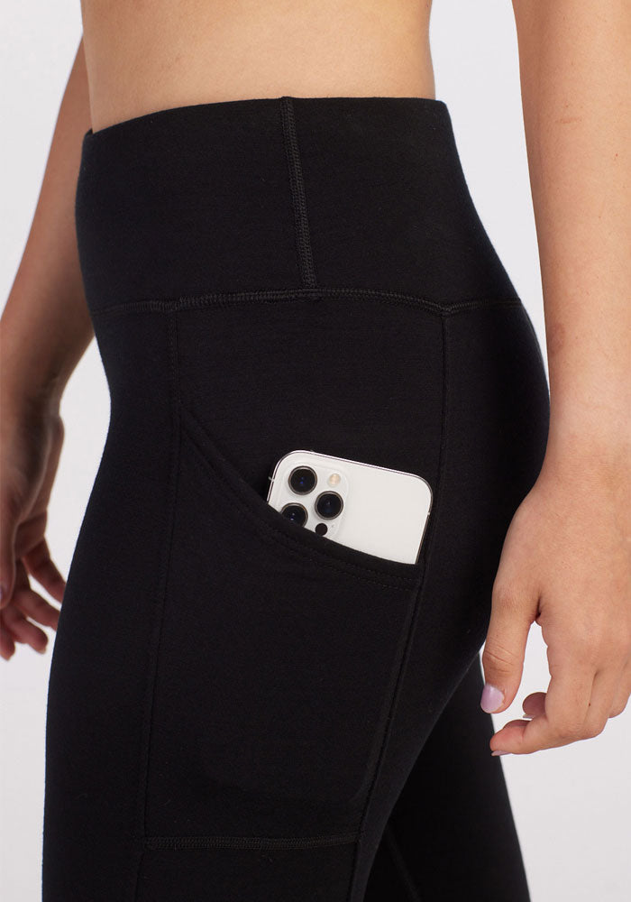 A close-up of a person wearing Woolx Piper Pocket Leggings in black, with a smartphone tucked into one of the side pockets. Only a portion of the person's torso and legs are visible against a plain, light grey background.