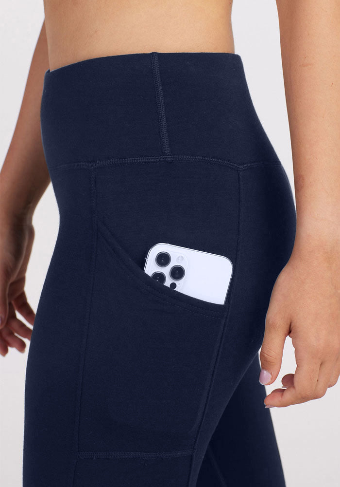 A close-up of a person wearing Woolx Piper Pocket Leggings in Deep Navy, with a smartphone partially visible in one of the side pockets. Crafted from Australian Merino Wool, these high-waisted leggings are both stylish and functional. The person is standing against a light neutral background, with only their midsection and part of the right arm visible.