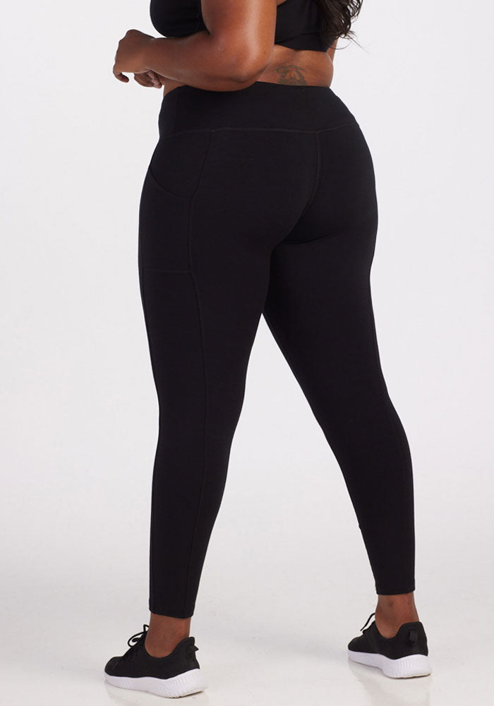 A person is standing facing away from the camera, wearing Woolx Piper Pocket Leggings in black along with a matching black top. Their hands are relaxed at their sides, and they are sporting black and white sneakers. The background is plain and neutral.