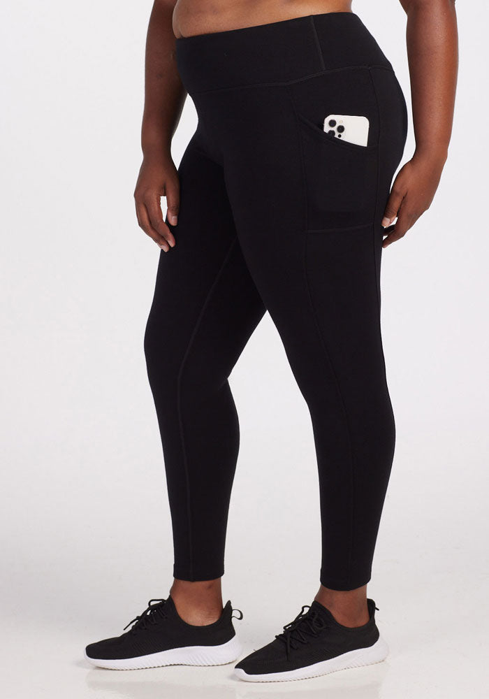 A person is dressed in Woolx's Piper Pocket Leggings in black, with a side pocket holding a white phone. They complete their look with black sneakers that feature white soles. Standing against a plain background, they maintain a relaxed posture. 