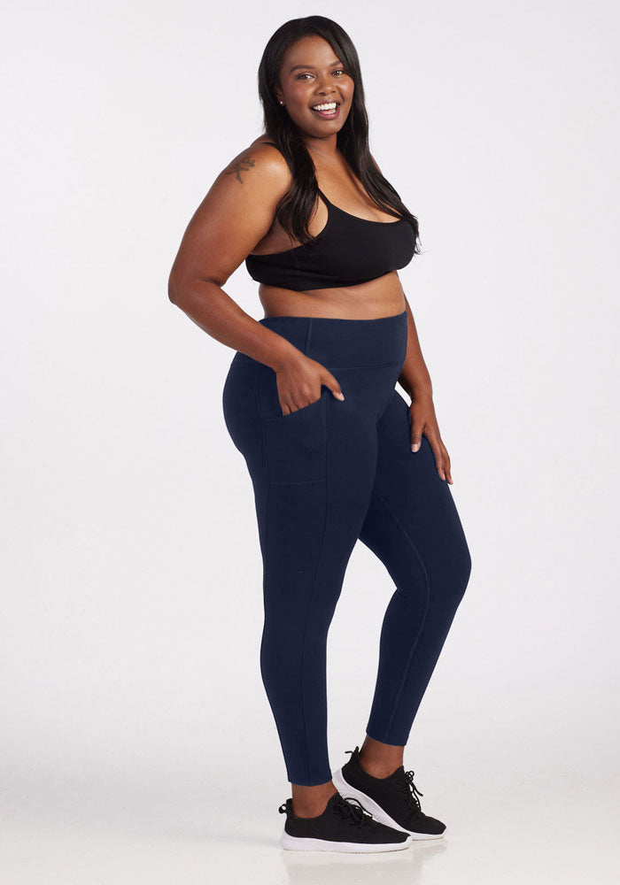 A woman poses confidently, wearing a black sports bra and Woolx's Piper Pocket Tall leggings in Deep Navy. She stands against a plain background, smiling and looking at the camera. Her long hair is down, and she completes her look with black sneakers.