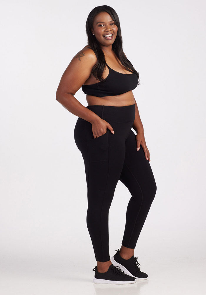 A woman with long, dark hair smiles while wearing a black sports bra, Woolx's Piper Pocket Tall - Black leggings featuring convenient pockets, and black sneakers with plush cushioning. She stands against a white background with her hands resting on the pockets of her leggings.