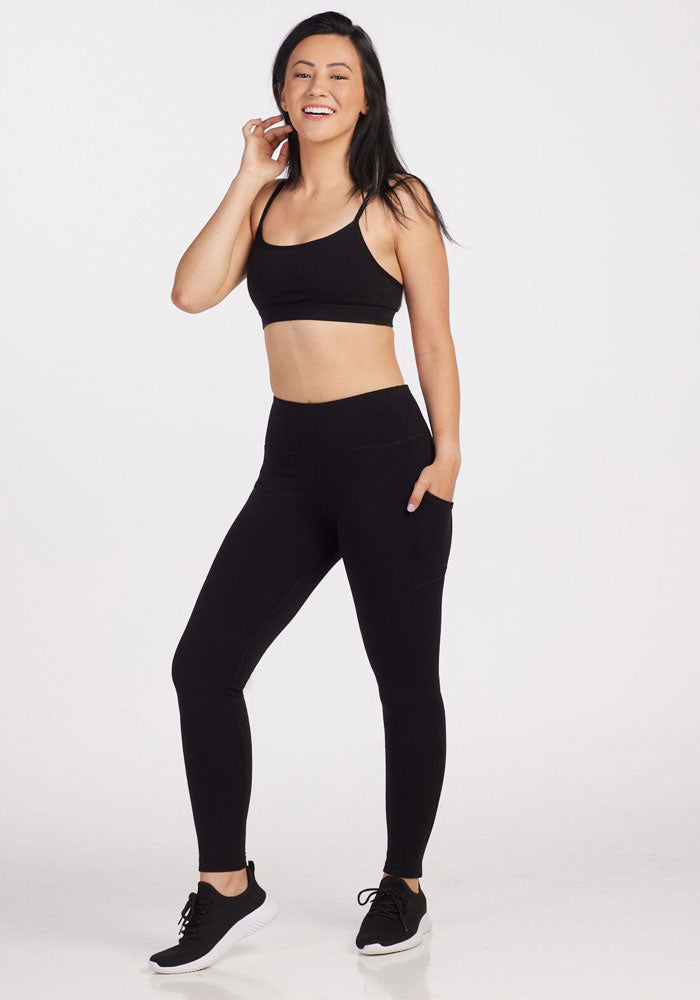 A woman stands smiling against a plain background, wearing a black sports bra, Piper Pocket Leggings - Black by Woolx, and black sneakers. She has one hand touching her face and the other resting in a pocket of her comfortable fit leggings.