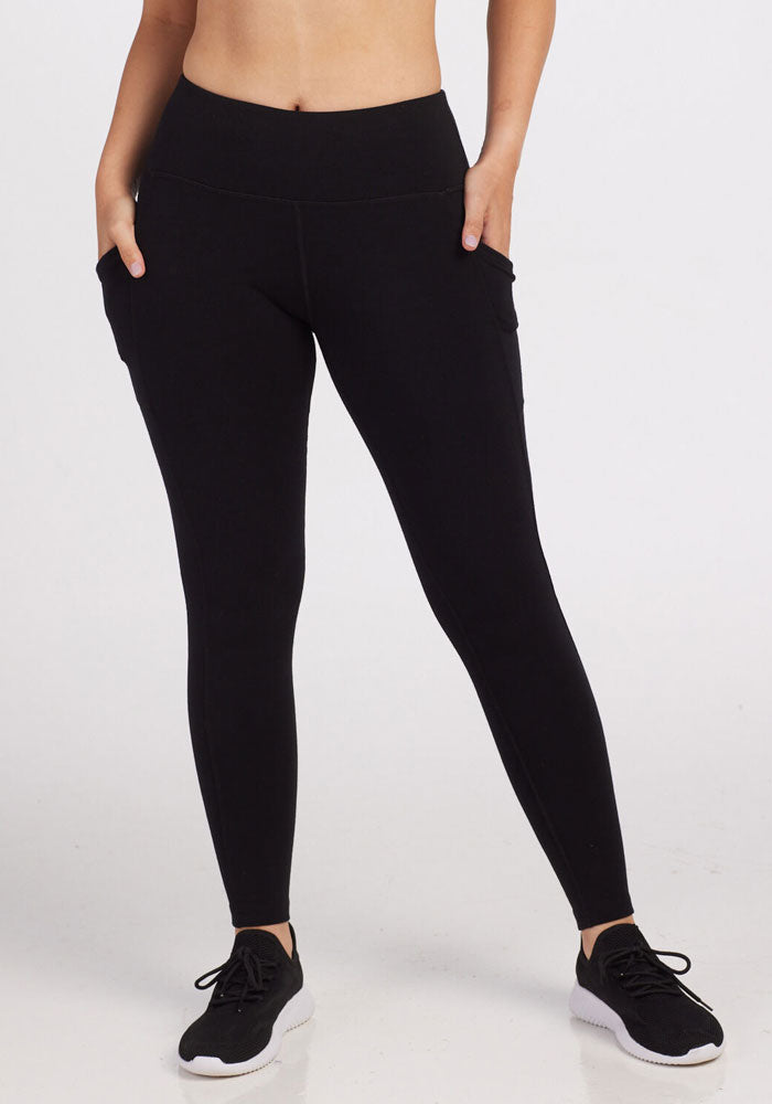 A person is wearing Woolx Piper Pocket Leggings in black with their hands in the side pockets. The form-fitting leggings are paired with black running shoes featuring white soles. Only the lower torso and legs of the person are visible against a plain white background. 