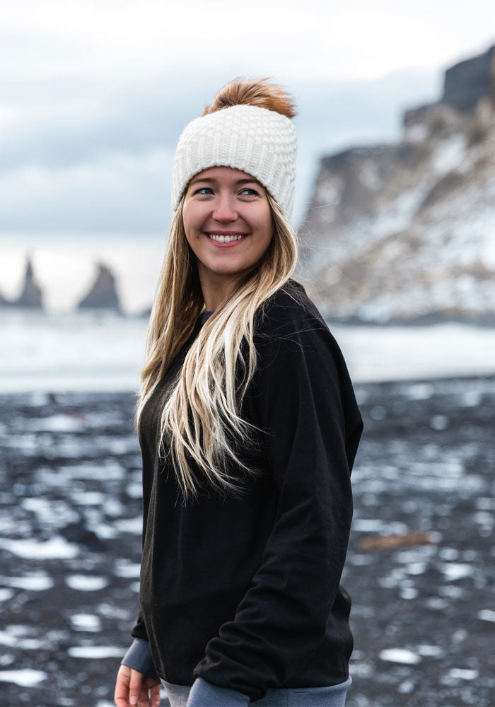 A smiling woman with long blonde hair dons a black sweater and the Woolx Kaylee Beanie in cream, featuring a chunky knit design with a faux fur pom-pom. She stands on a rocky shoreline, where patches of snow sprinkle the scene and blurred cliffs rise in the background.