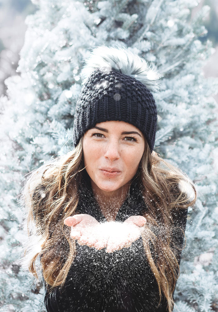 A woman donning the Woolx Kaylee Beanie - Black, featuring a chunky Merino wool knit and a faux fur pom-pom, stands in front of a snow-draped tree. She playfully blows snowflakes from her hands toward the camera, set against a bright and wintry backdrop.