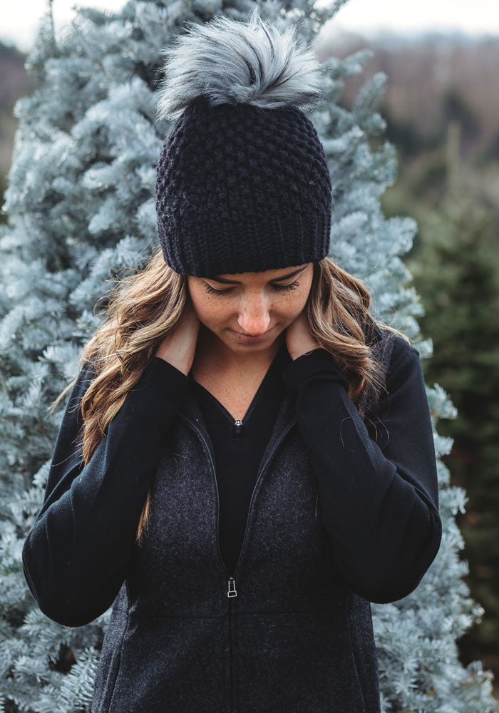 A woman wearing the Woolx Kaylee Beanie in black, featuring a chunky knit design and a faux fur pom-pom, stands outdoors before a frosted evergreen tree. She is gazing down while dressed in a black zip-up jacket.