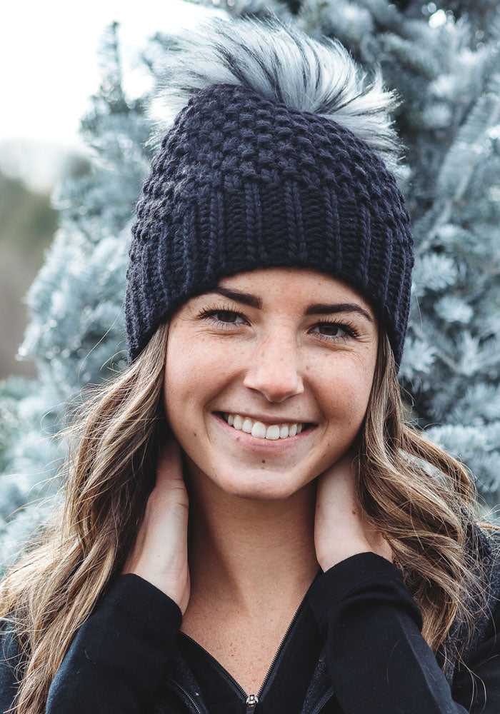 A woman smiles, adorned with the Woolx Kaylee Beanie in black, featuring a gray faux fur pom-pom. She stands before a frosty evergreen tree, hands near her neck, dressed in a black top.