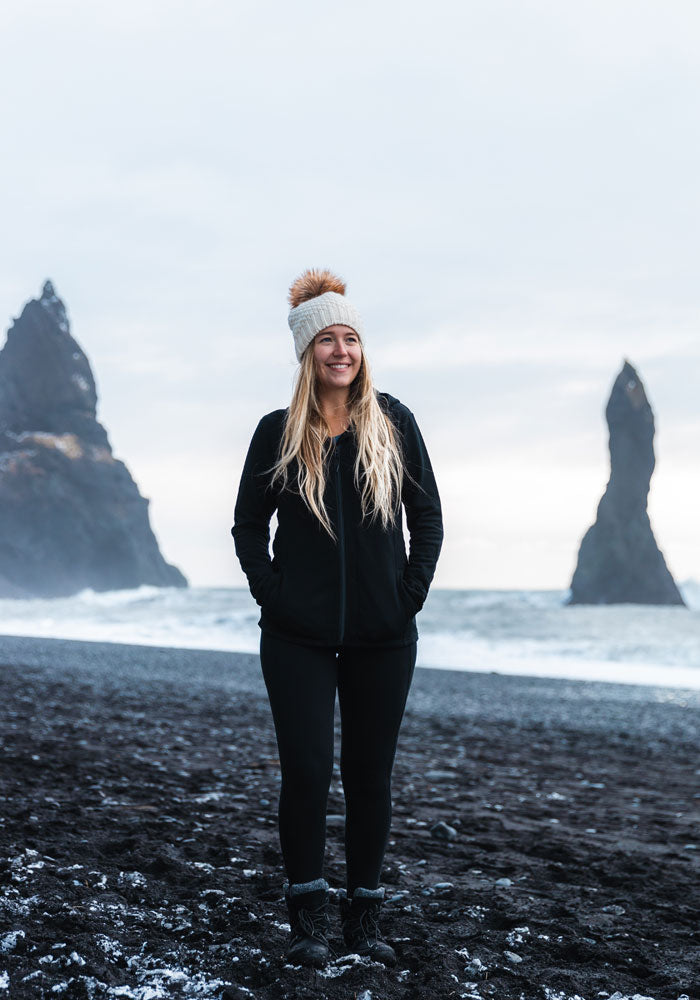 A person stands smiling on a black sand beach, wearing the Kaylee Beanie in cream by Woolx and a black jacket. The ocean and large rock formations are in the background under a cloudy sky.