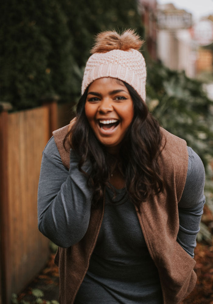 A person with long hair, wearing a chunky knit Kaylee Beanie in rose by Woolx with a faux fur pom-pom and a brown vest over a gray shirt, is leaning forward and laughing joyfully. The background features greenery and a wooden fence.