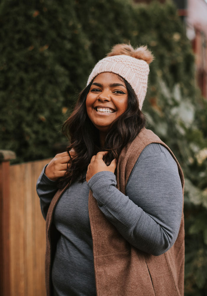 A woman smiling warmly at the camera, wearing the cozy Kaylee Beanie in rose by Woolx, featuring a faux fur pom-pom, paired with a brown vest over a gray long-sleeve shirt. She stands outdoors with greenery in the background.