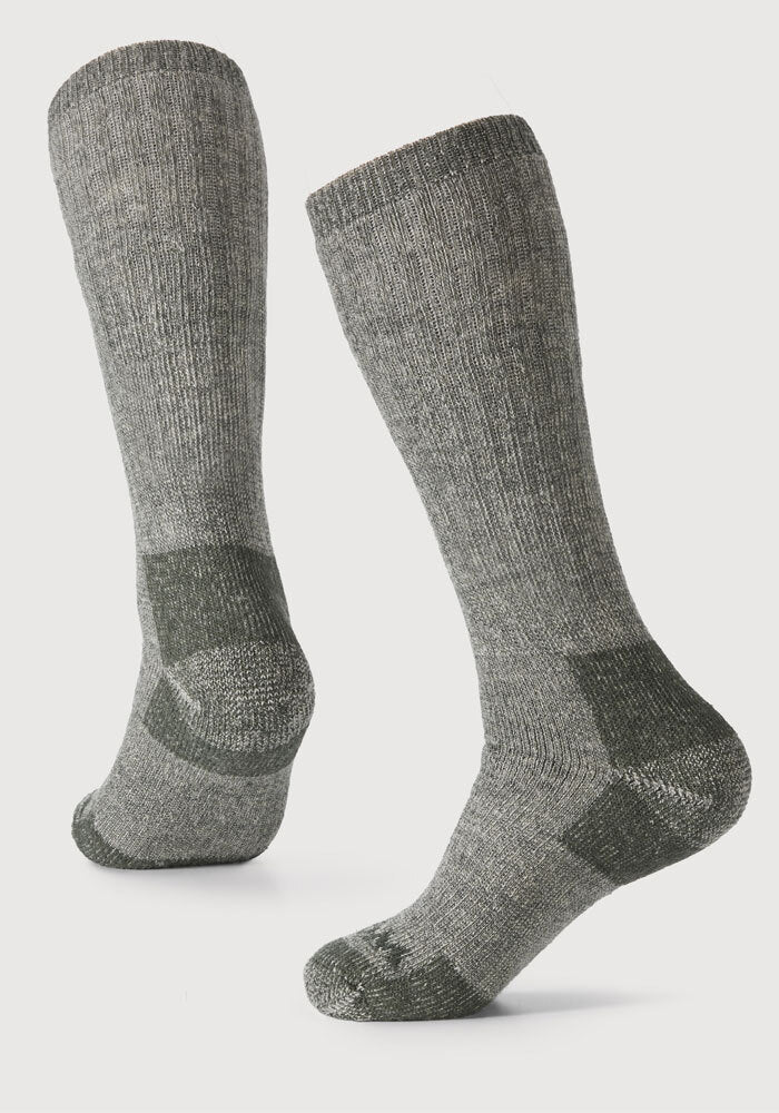 Against a plain background are two dark moss over-the-calf socks from Woolx, featuring reinforced heels and toes. These socks are made from heavyweight merino wool with a ribbed texture, offering a cozy, moisture-wicking, and durable look.